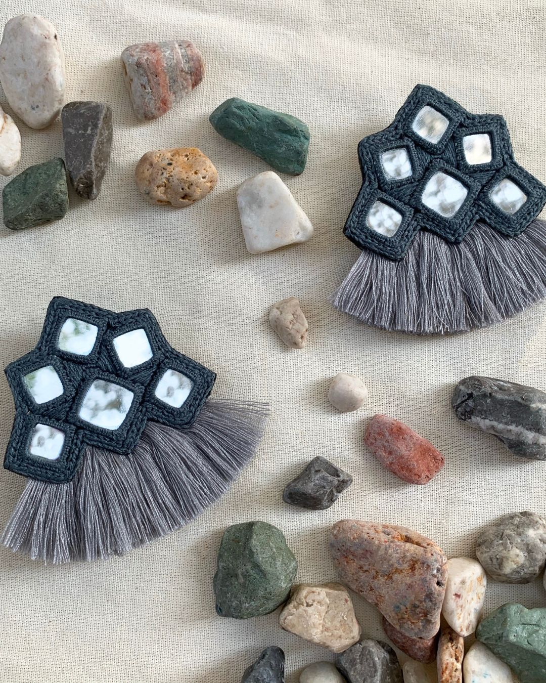 Handmade black macramé tassel earrings with white square stone accents and soft gray fringe, styled on natural linen fabric with decorative pebbles for a modern boho jewelry flat lay.