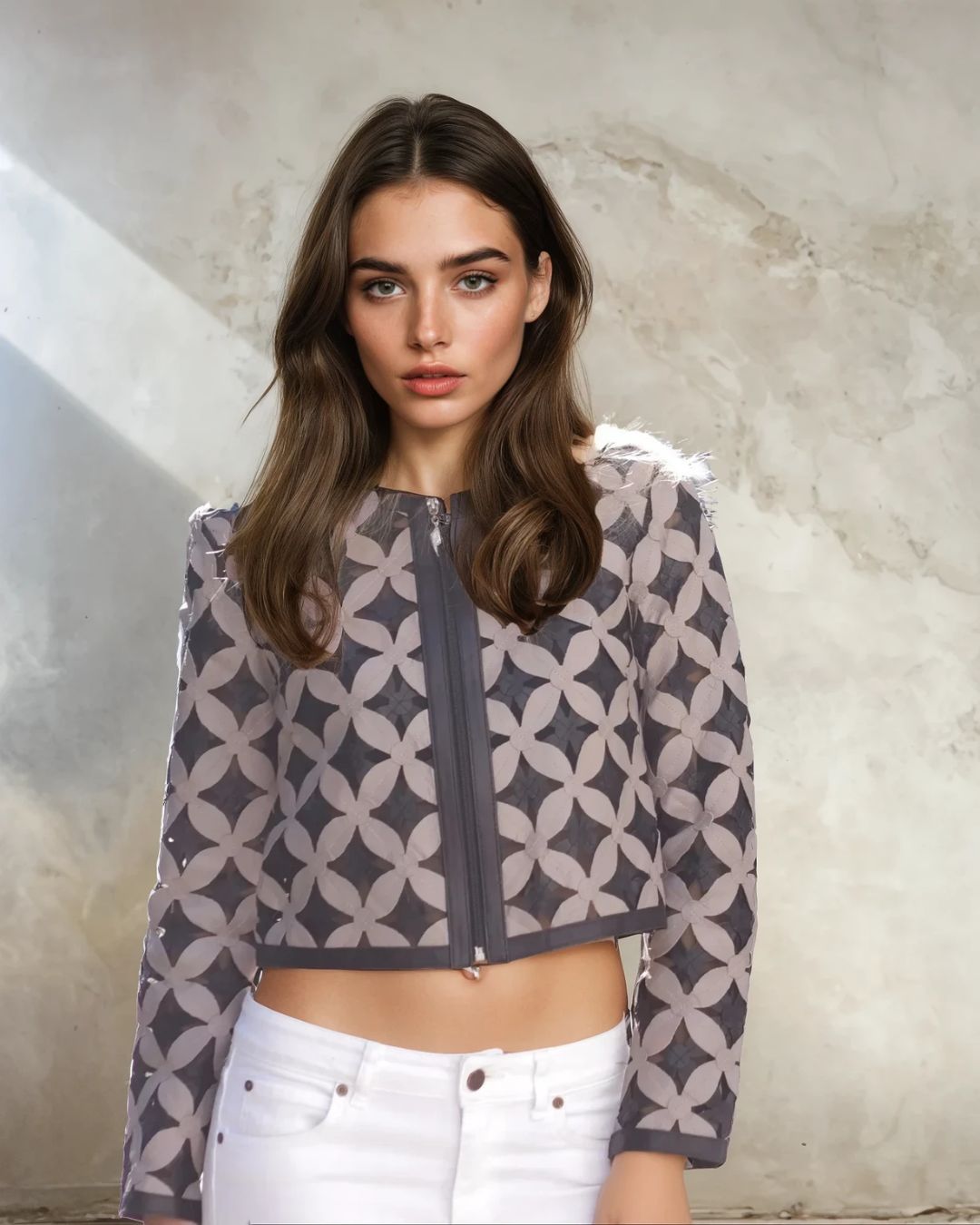 Woman wearing a patterned jacket and white pants against a textured wall.