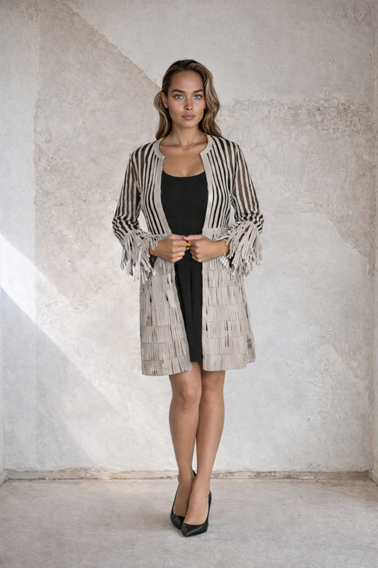 Woman wearing an elegant beige and black striped sheer longline jacket with fringe detailing over a black dress, styled with black heels in a modern minimalist setting.