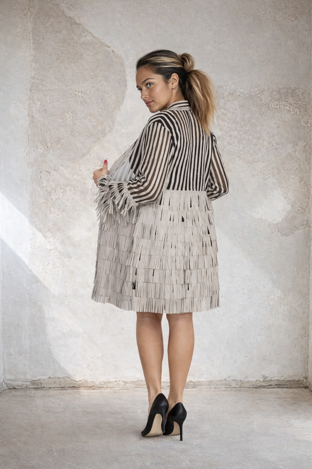 Back view of a woman wearing an elegant beige and black striped sheer longline jacket with layered fringe detailing and long sleeves, paired with a black dress and black high heels in a modern minimalist setting.