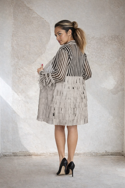 Back view of a woman wearing an elegant beige and black striped sheer longline jacket with layered fringe detailing and long sleeves, paired with a black dress and black high heels in a modern minimalist setting.