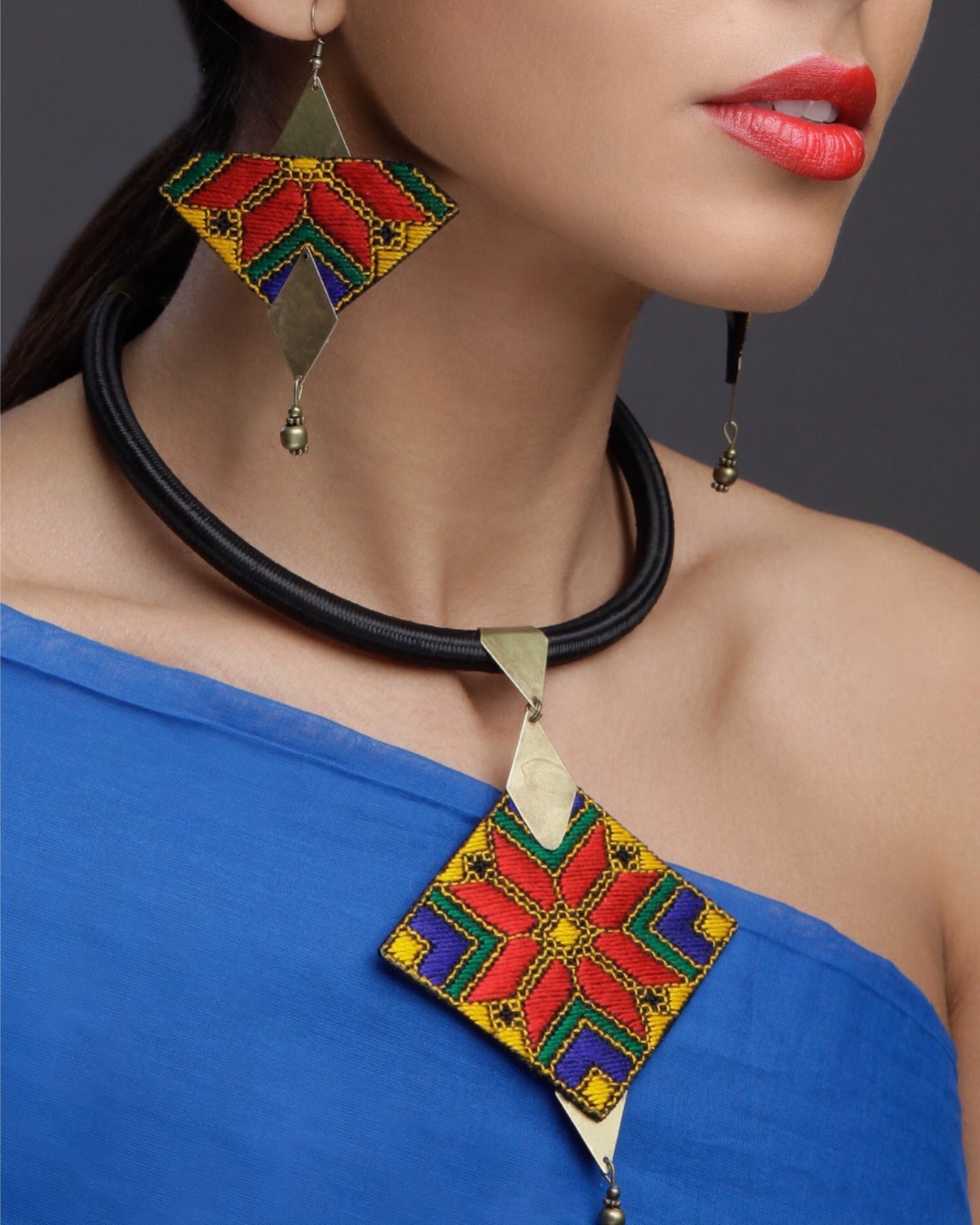 Close-up of a woman wearing a handcrafted geometric jewelry set featuring colorful embroidered diamond-shaped statement earrings and a matching pendant necklace with brass accents in vibrant red, yellow, blue, and green patterns.